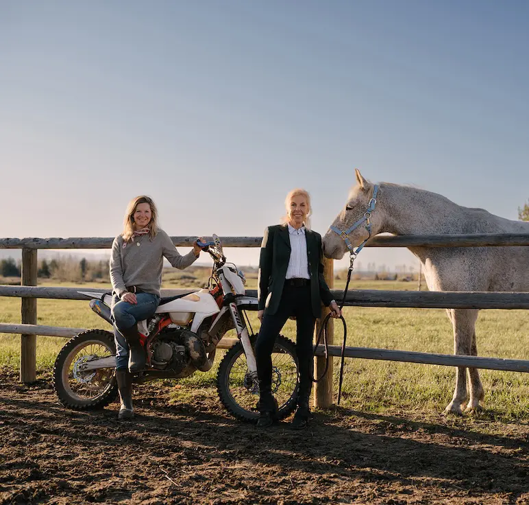 Ouest House Founders Anna Cole and Jenny Walton in front of wooden ranch fence, with Anna on a dirt bike and Jenny next to a horse.